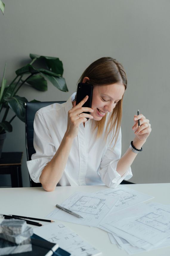 Woman in white shirt on the phone, looking at architectural drawings at a desk, smiling.