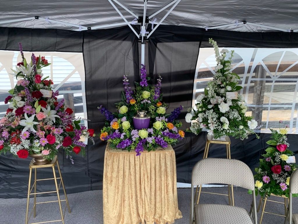 Floral memorial display under a tent with three flower arrangements and two empty chairs