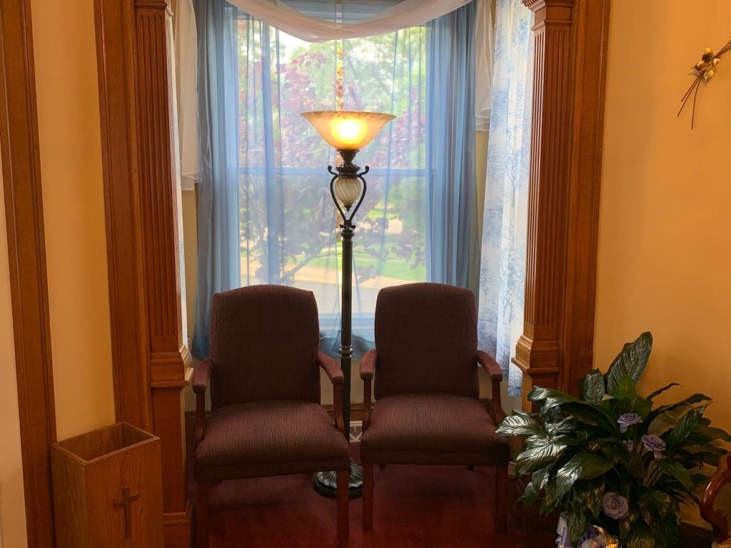 Two brown chairs by a window with sheer curtains, a lit floor lamp, and a potted plant in a cozy room