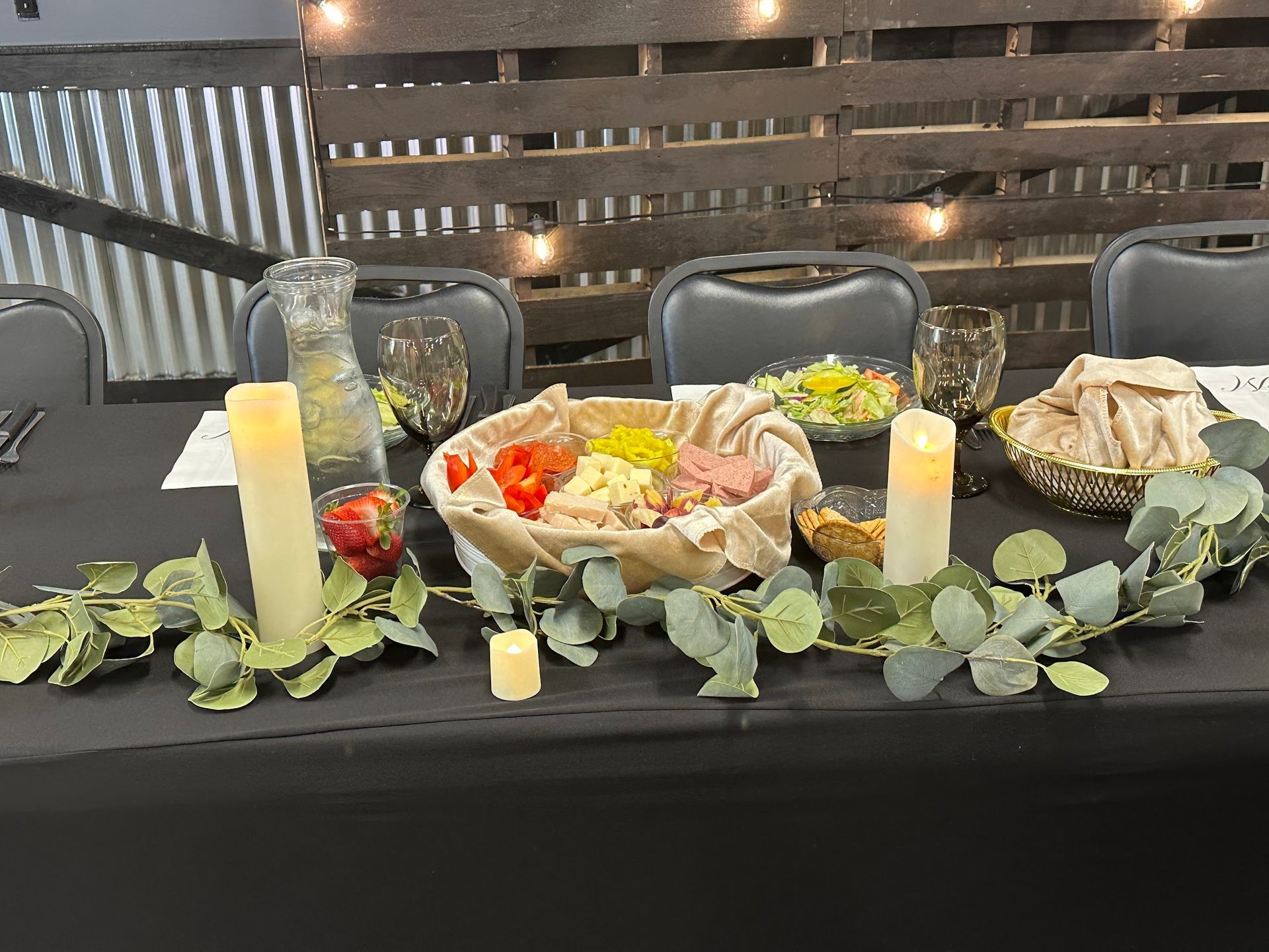 Buffet table with platters of sandwiches, candles, eucalyptus decor, and string lights against a striped backdrop