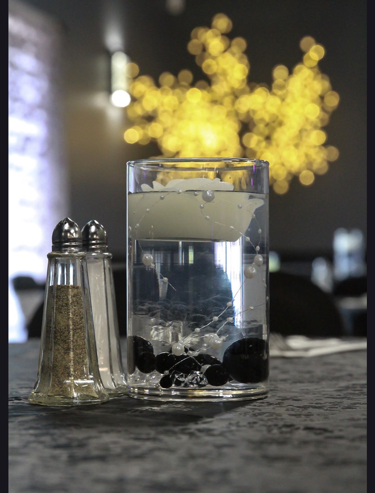 Cocktail glass with ice and black berries on a table, with a salt shaker and warm bokeh lights behind it