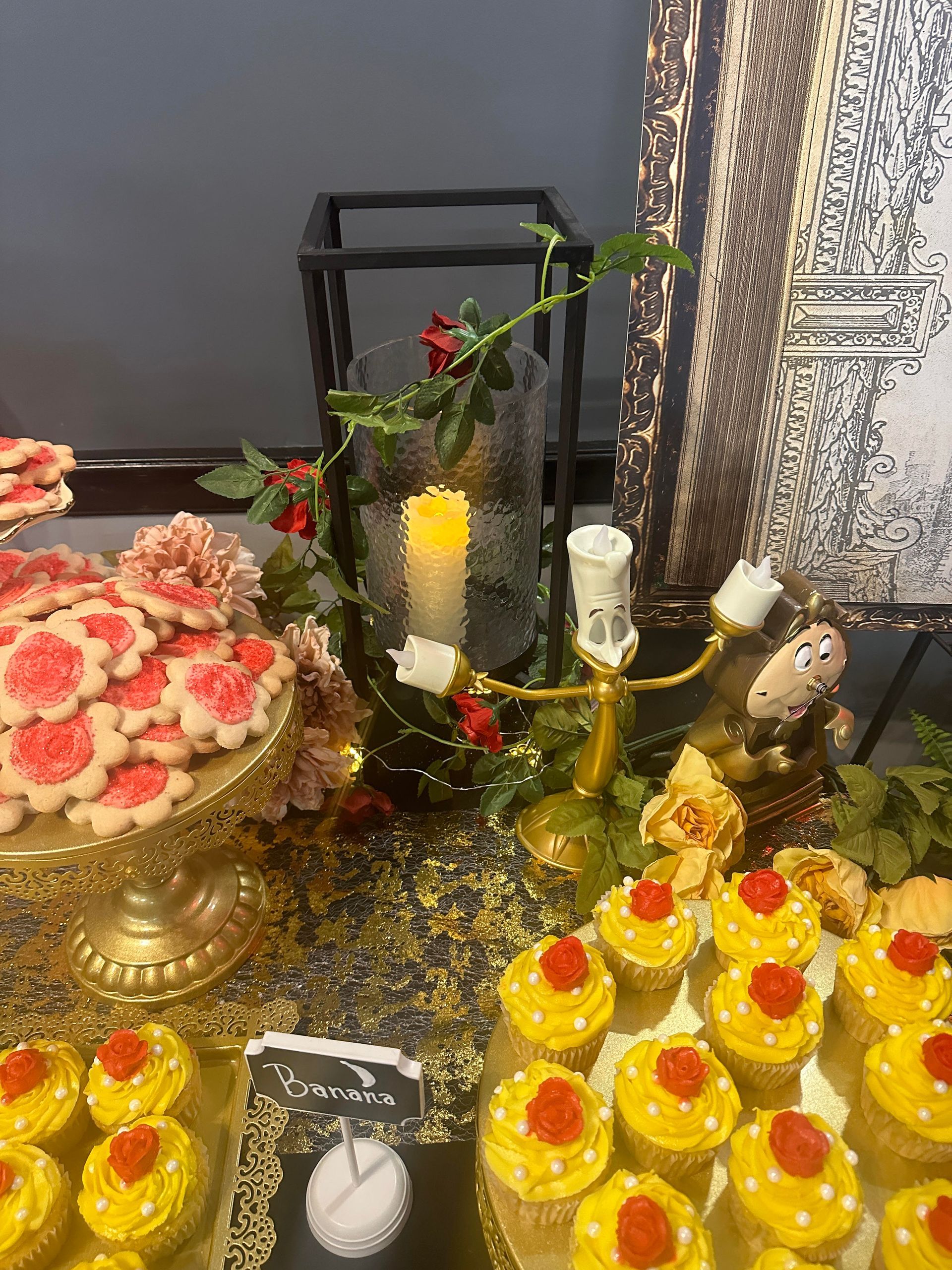 Dessert table with gold trays of yellow sweets, red floral cake, and candles on a glittery display