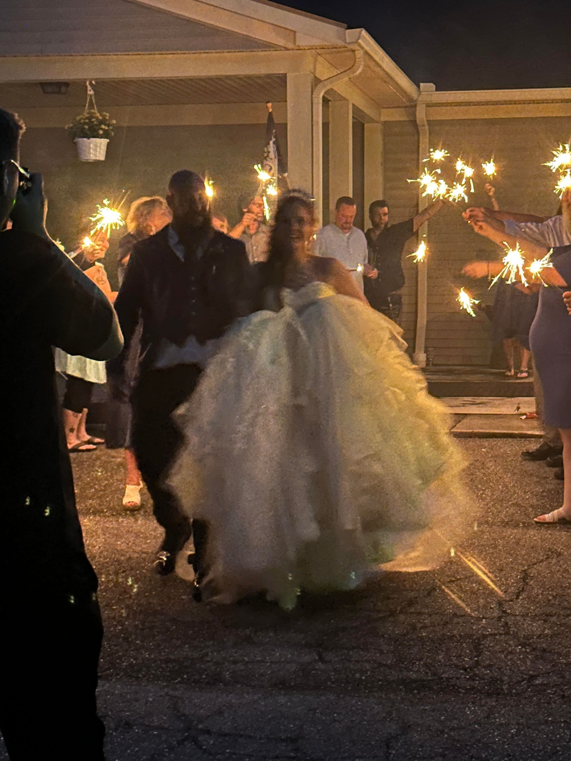 Couple in formal attire walks through sparkler-lit outdoor wedding reception at night