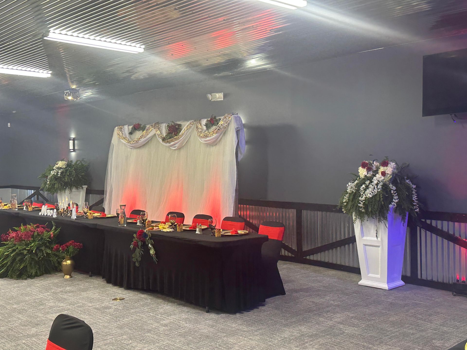 Decorated event hall with a black-draped stage table, red lighting, and white floral arrangements