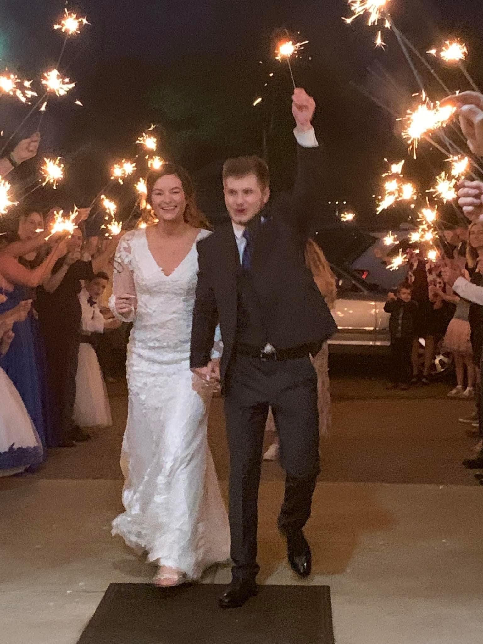 Wedding couple walking down a sparkler-lit aisle, the bride in a white gown and the groom in a dark suit.
