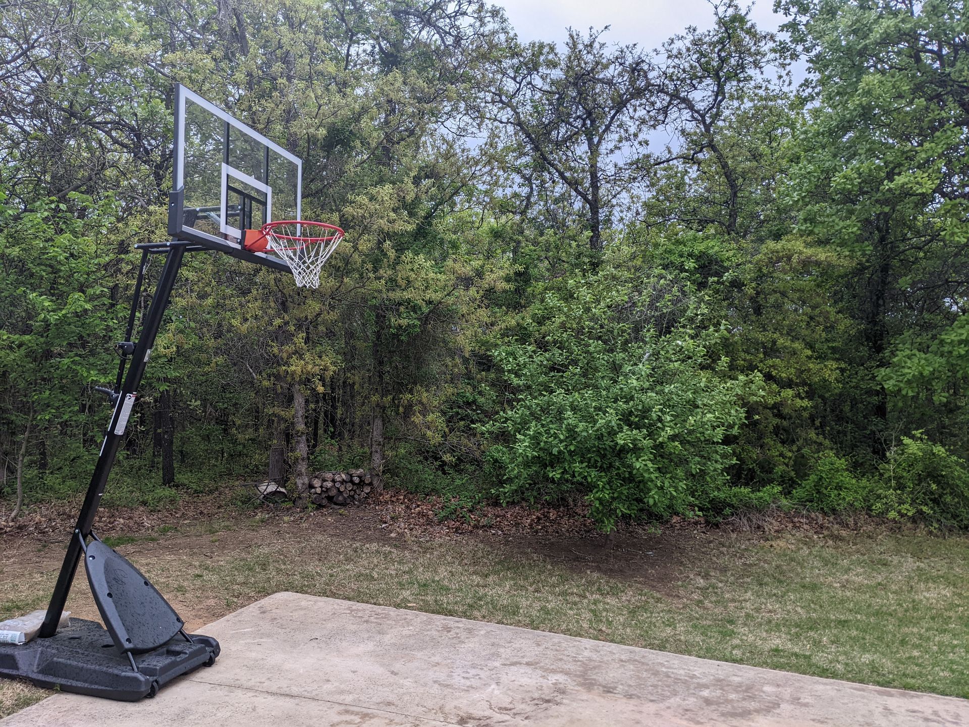 Portable basketball hoop on a concrete pad beside a grassy yard and trees.