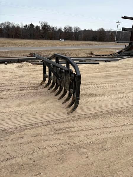 Black metal rake attachment on a tractor in a dirt field near a wooded edge