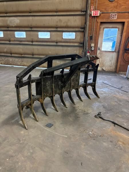 Large black metal grapple attachment with curved tines in a workshop garage.