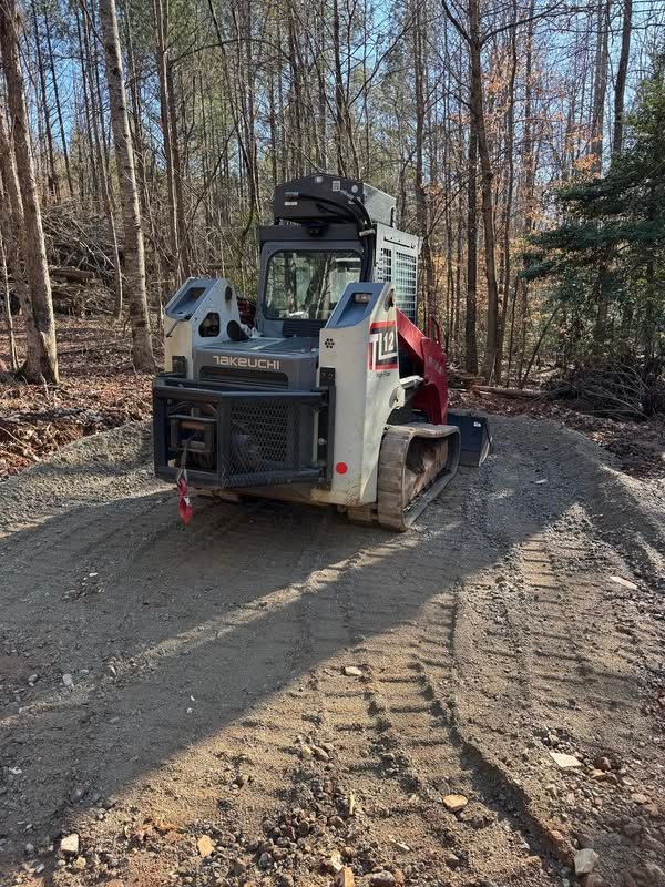 Tracked skid steer on a muddy forest road, carrying a person in a red jacket during fall.