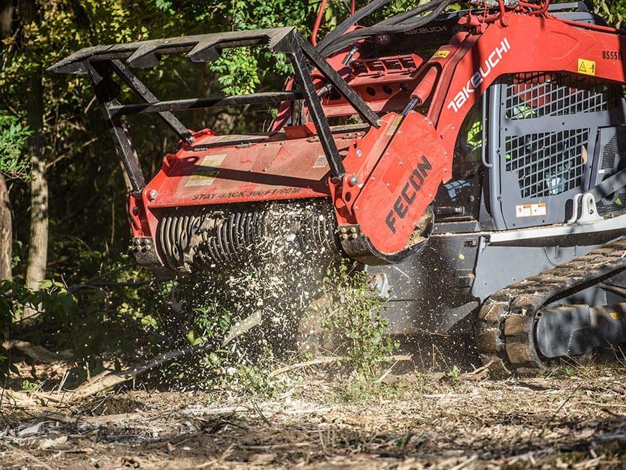Red skid steer clearing brush, spraying debris in a wooded area