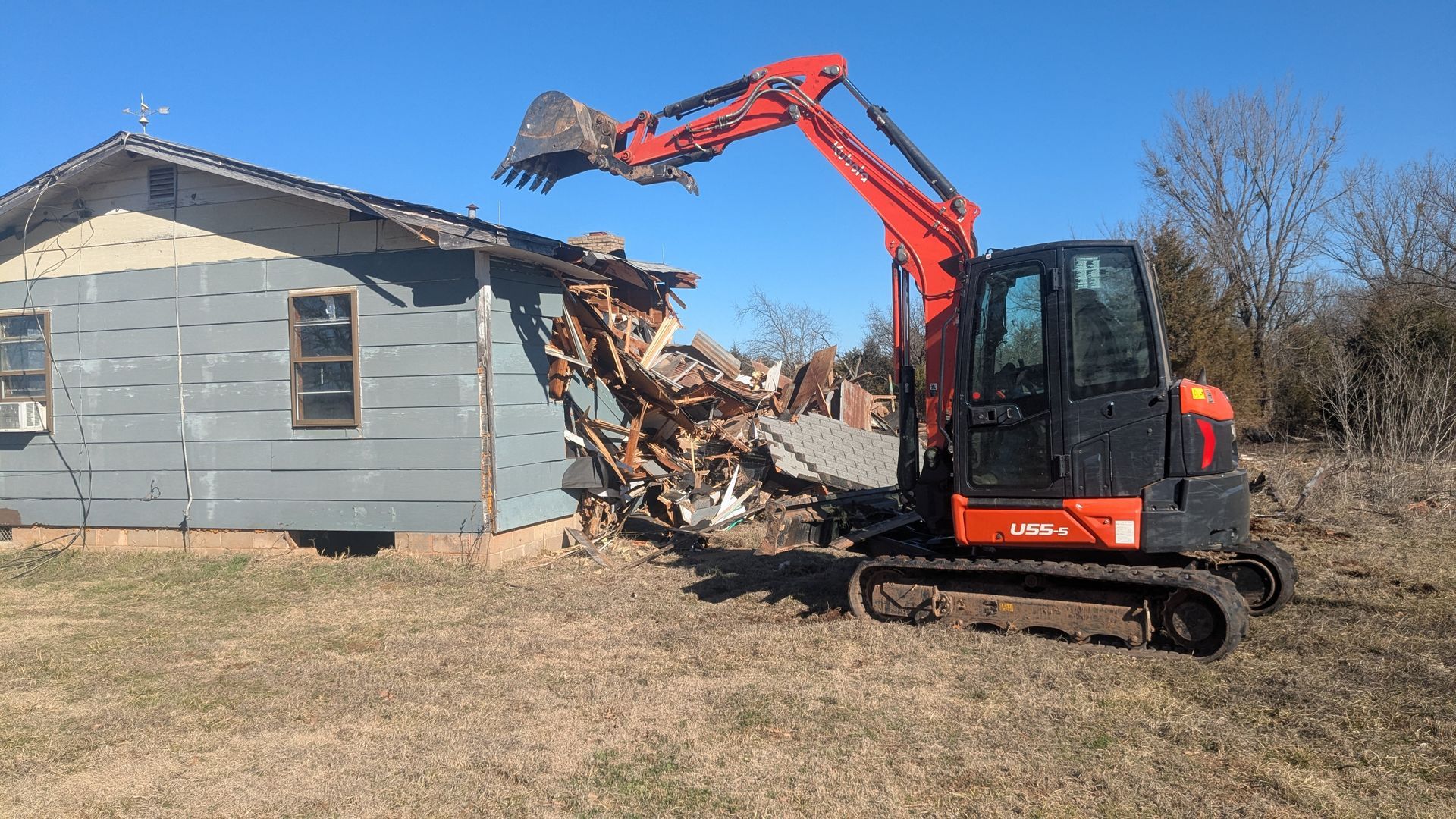Orange excavator demolishing a gray house in a rural yard