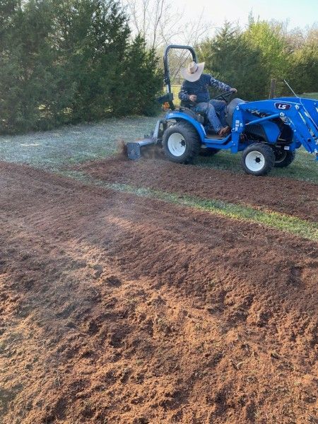 Blue tractor tilling a muddy field beside a frosty tree line at sunrise