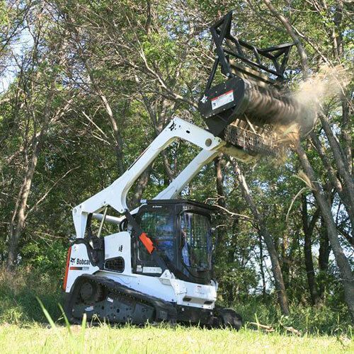 Compact tracked loader with brush cutter clearing trees in a grassy woodland area.