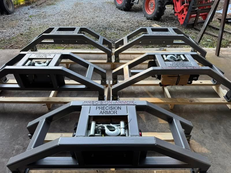 Three black metal mower frames on wooden pallets outdoors, with a tractor in the background