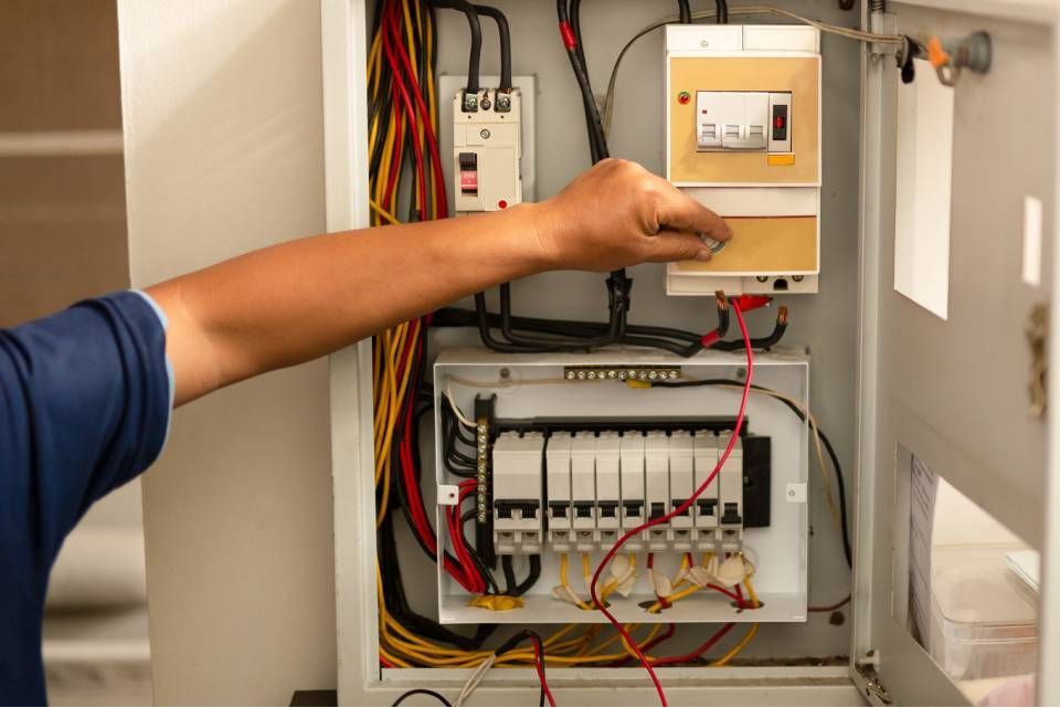 Arm pointing at an electrical panel with exposed wires inside a beige utility cabinet
