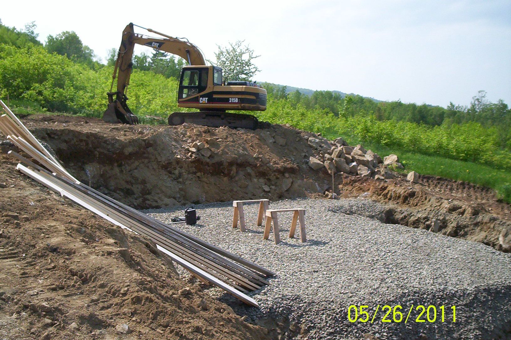 Excavator at a dirt excavation site with gravel, pipes, and a date stamp in the corner