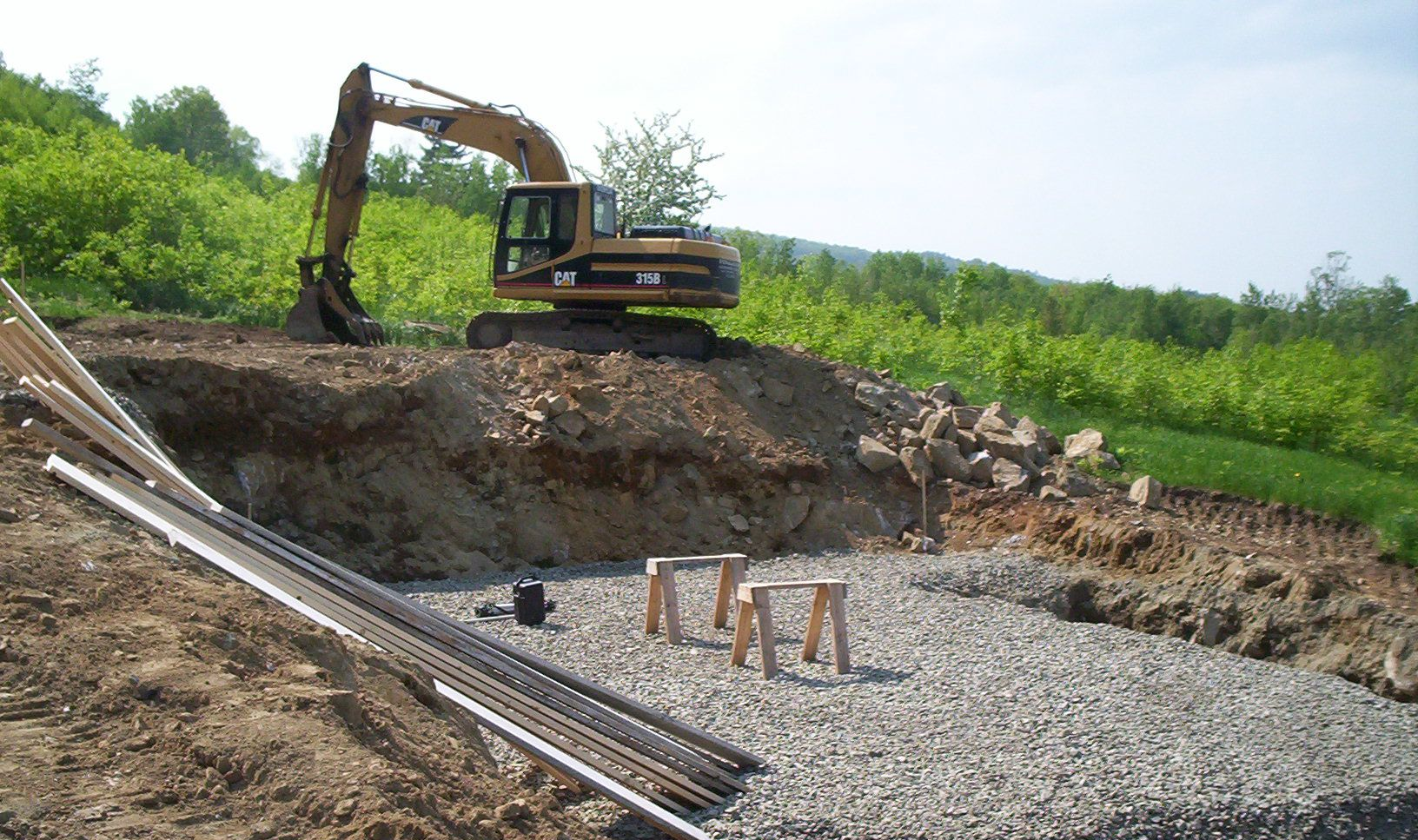Excavator digging a large gravel pit beside piles of pipes and green trees under a cloudy sky