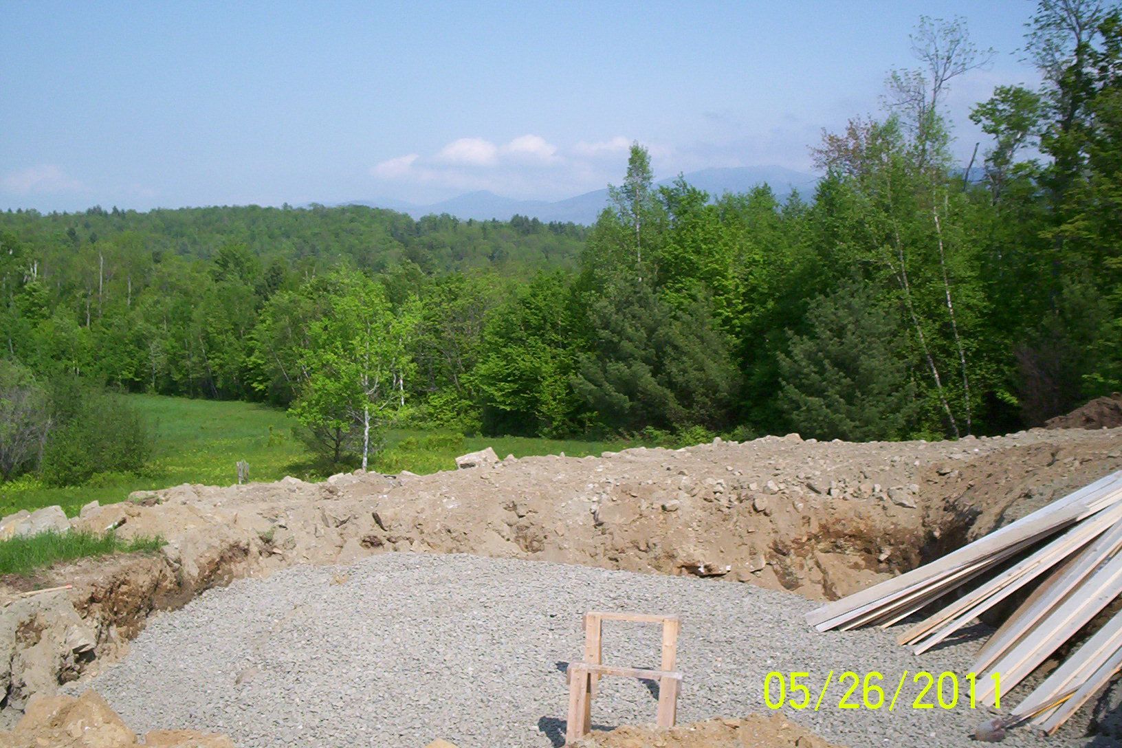 Gravel excavation site with dirt berms, pipes, and a green wooded hillside under a blue sky