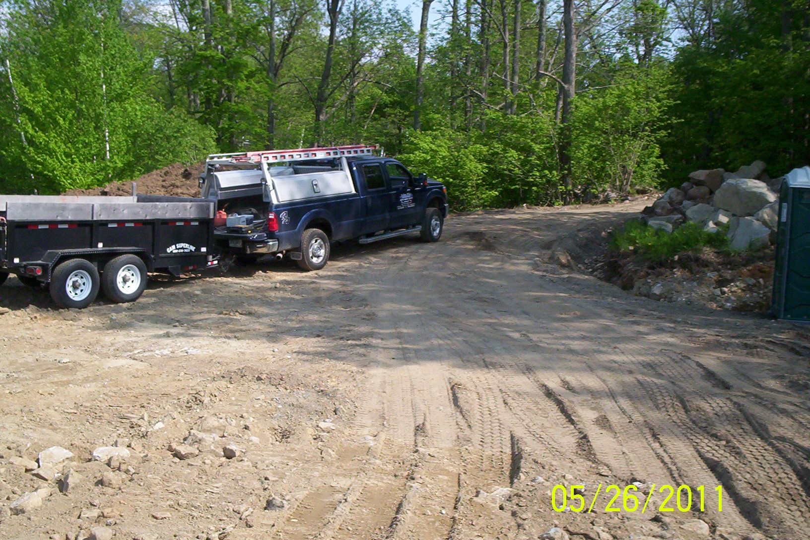 Black pickup truck towing a flatbed trailer on a dirt road beside trees and rocks