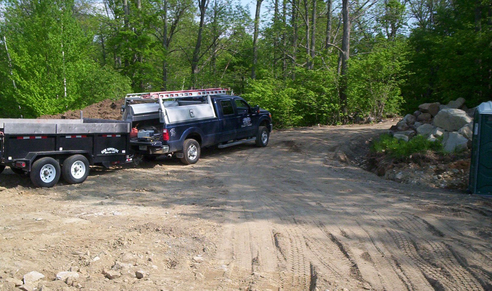 Black pickup truck and trailer parked on a dirt clearing beside a wooded area.