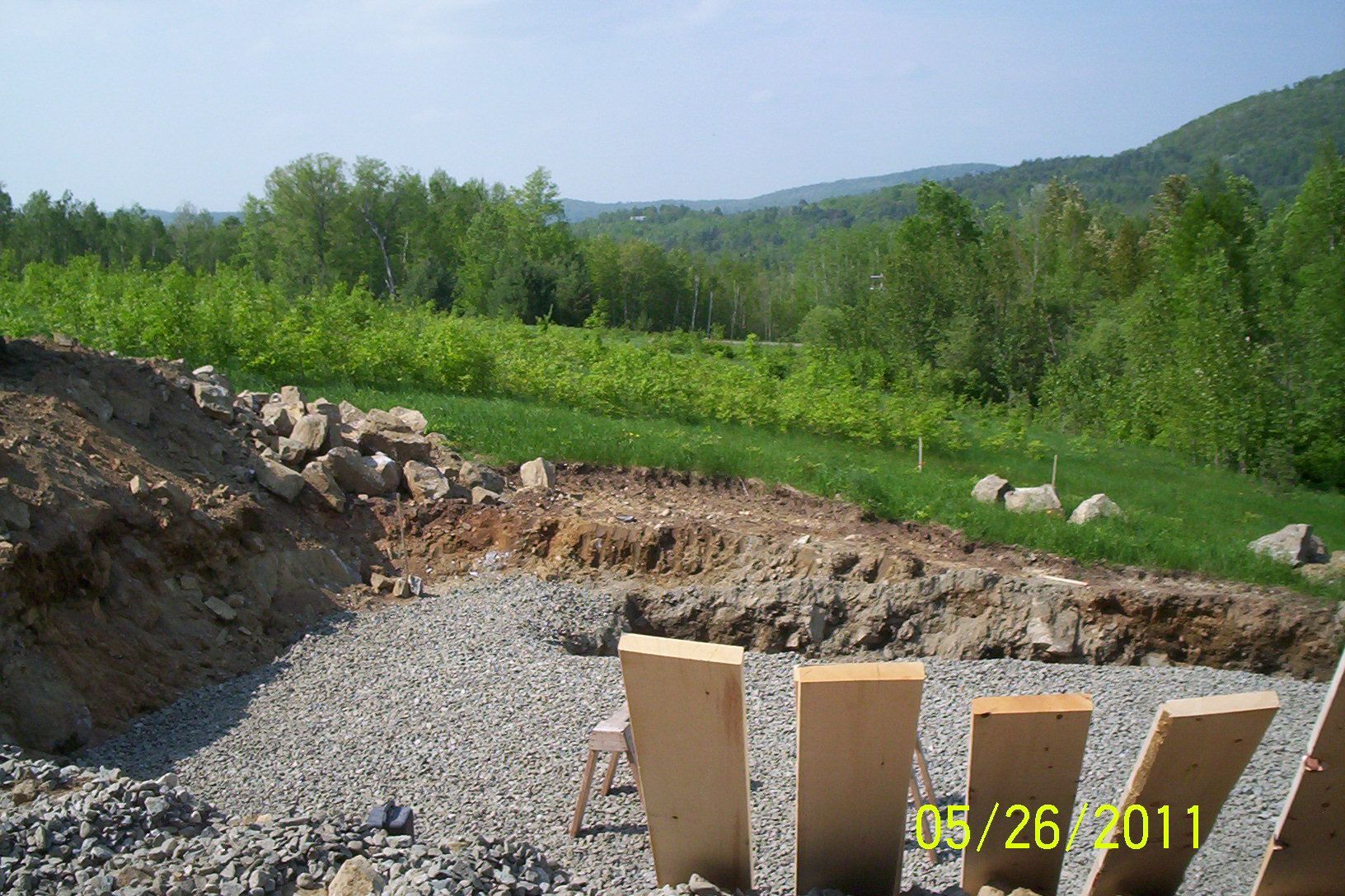 Gravel construction site with dirt excavation, wooden forms, and green hills in the background