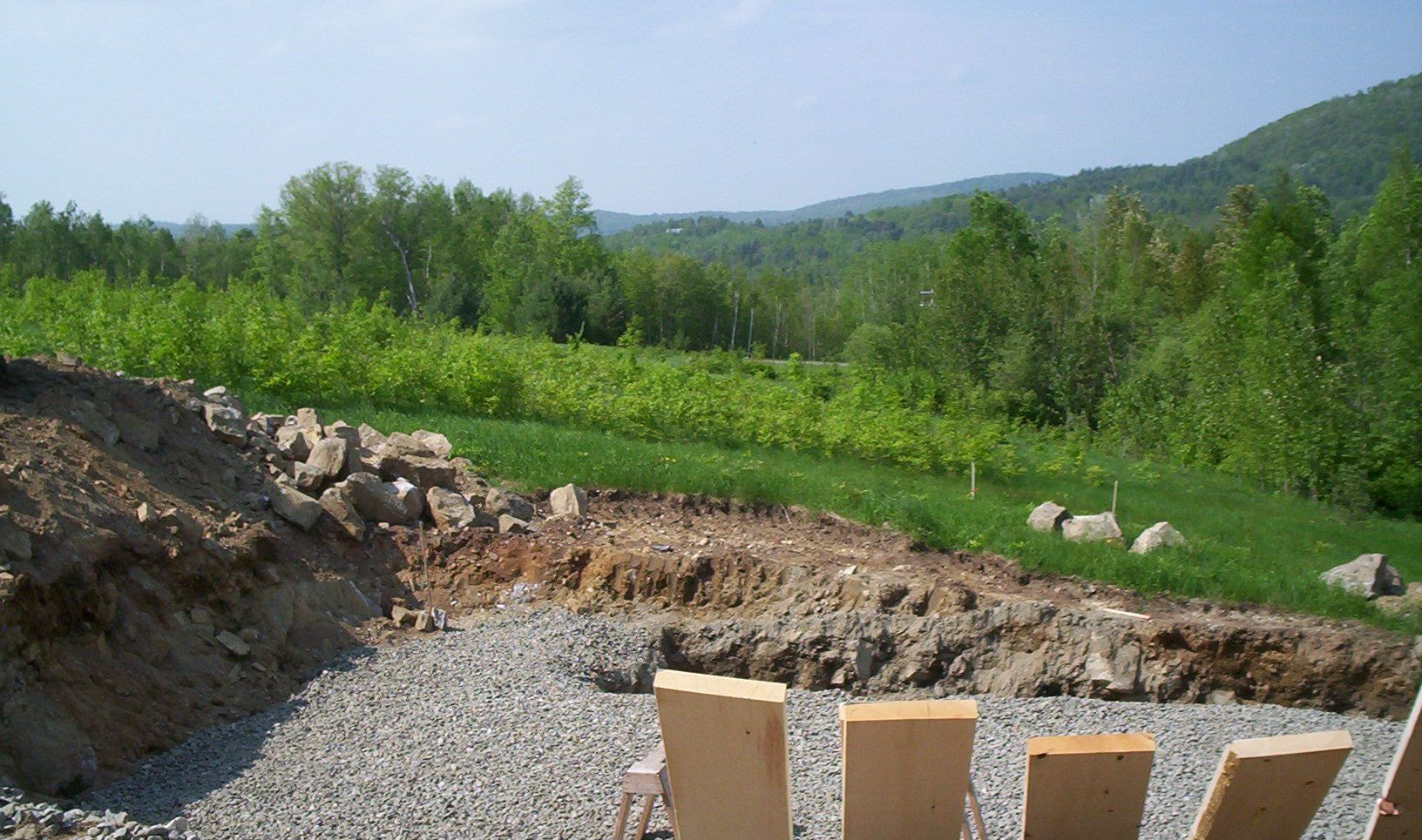 Gravel construction site overlooking a green valley with trees and hills under a clear sky