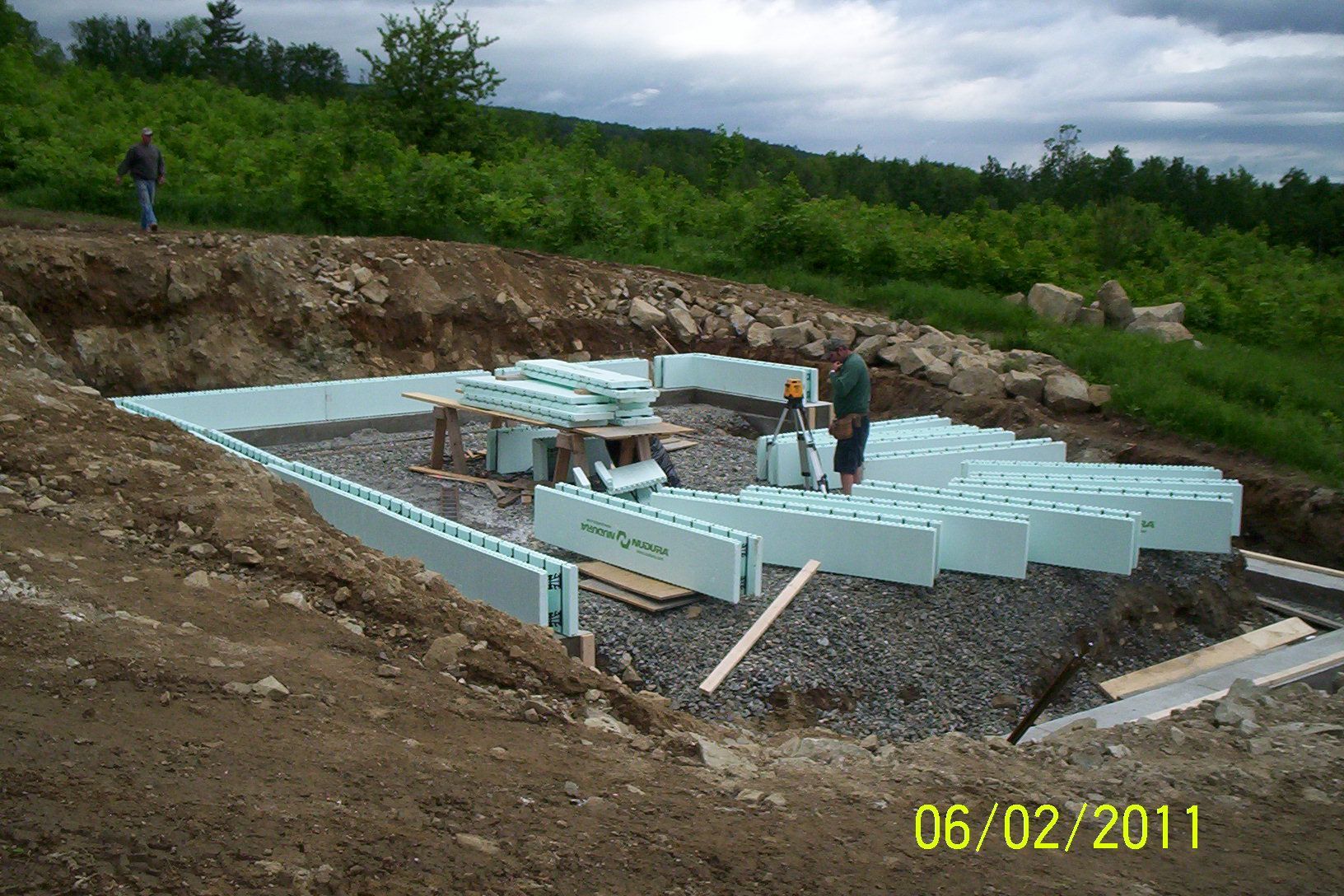 Workers build a light-blue concrete foundation in an excavated site surrounded by dirt and greenery.