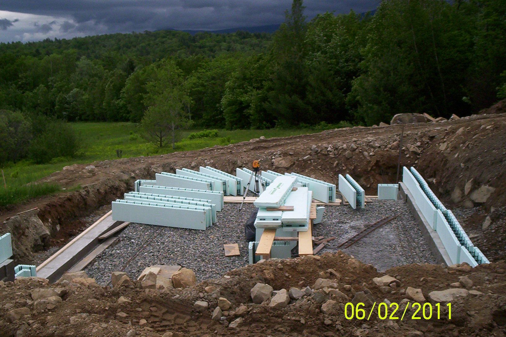 Construction site with white foam blocks and lumber on gravel, surrounded by trees and dirt under cloudy sky