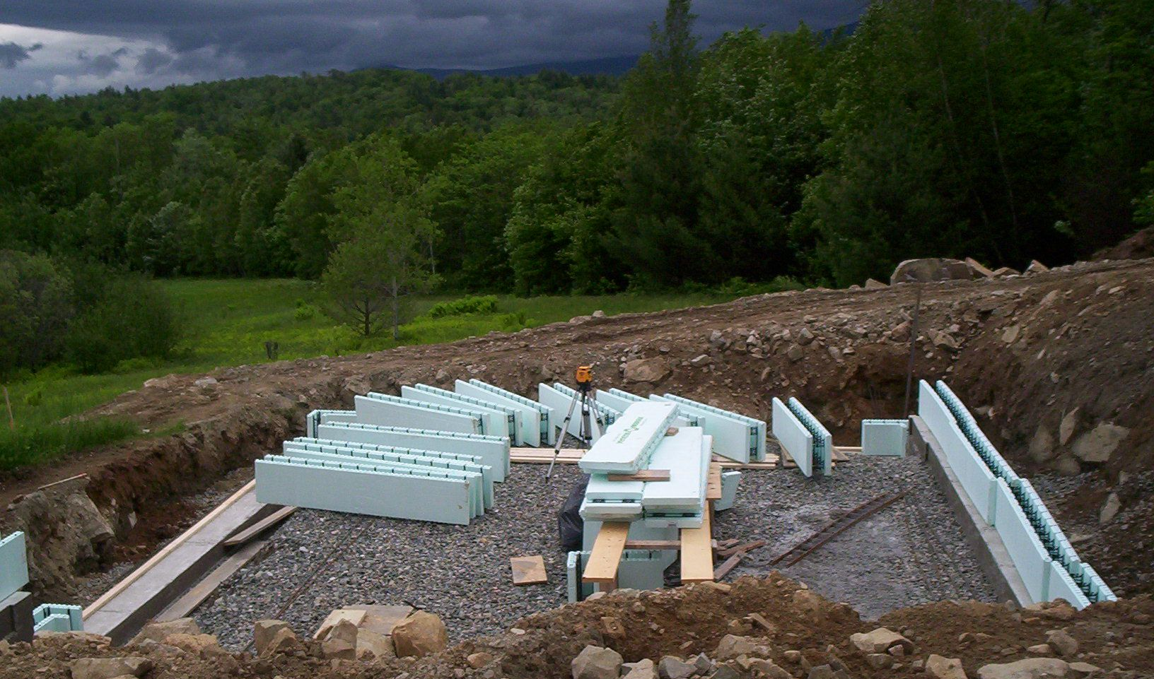 Partially built foundation with light blue insulation blocks on a gravel site beside a wooded hillside