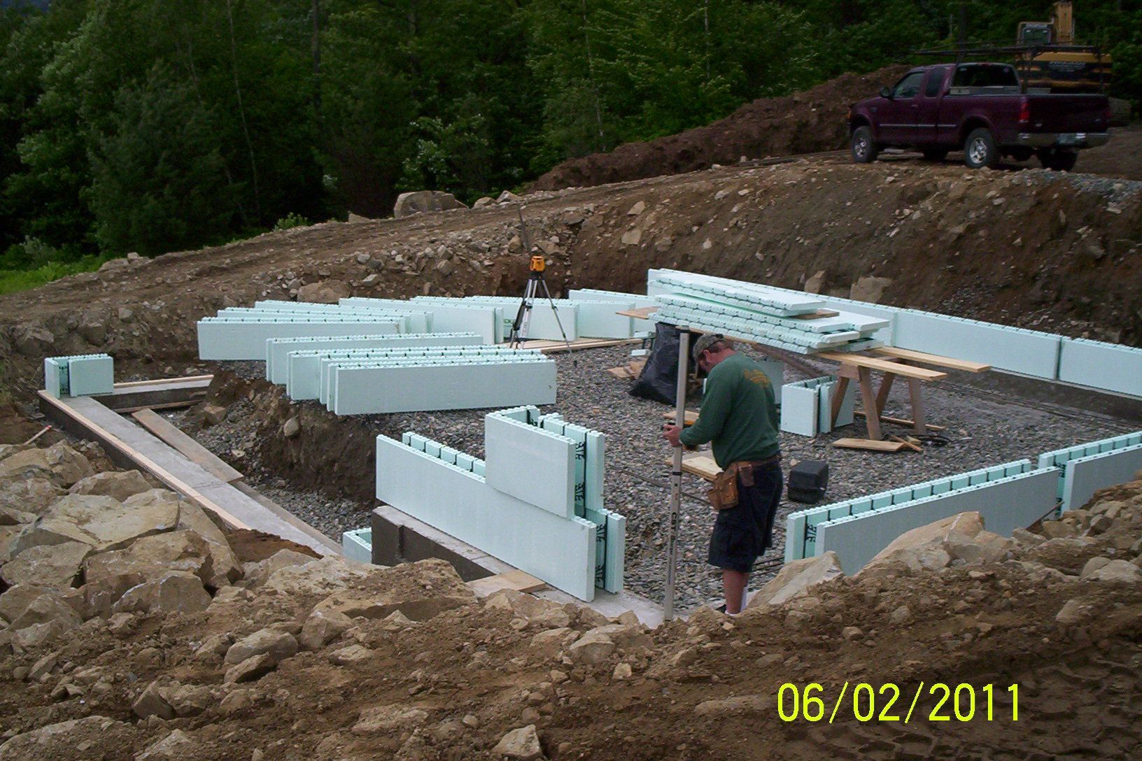 Foundation walls under construction on a dirt lot, with workers assembling white insulated blocks.