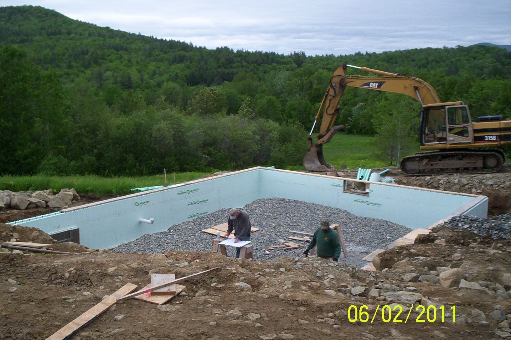 Excavator at a concrete foundation site in a wooded rural area with two workers nearby.