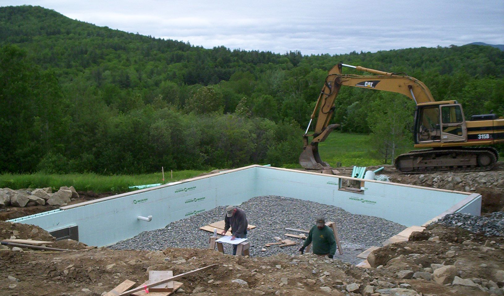 Excavator digging a rectangular pool foundation at a rural construction site with gravel and wooded hills.