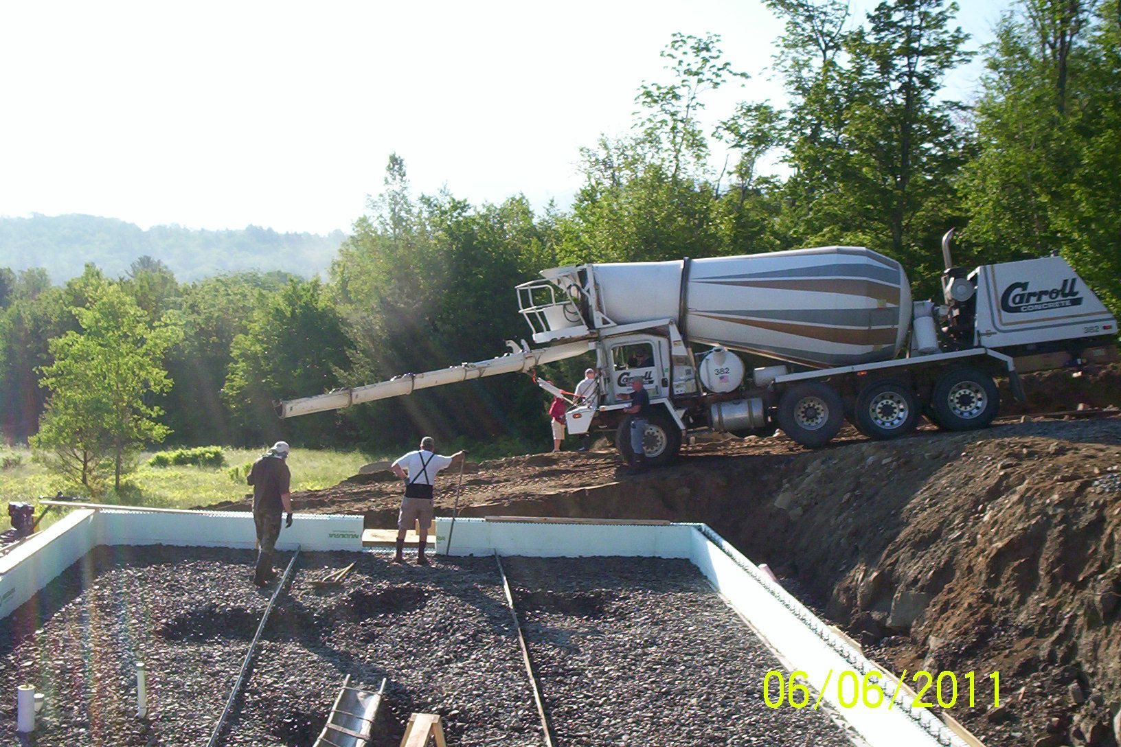 Concrete mixer truck pouring concrete into a foundation on a gravel construction site with two workers nearby