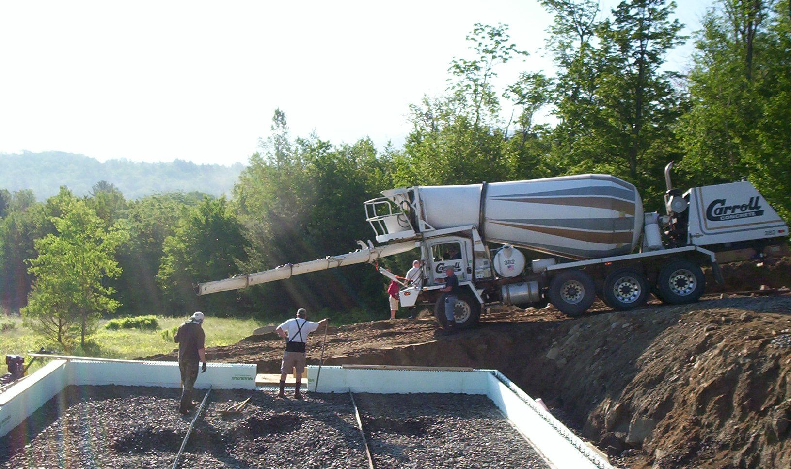 Concrete truck pouring into a foundation pit at a rural construction site, with workers nearby