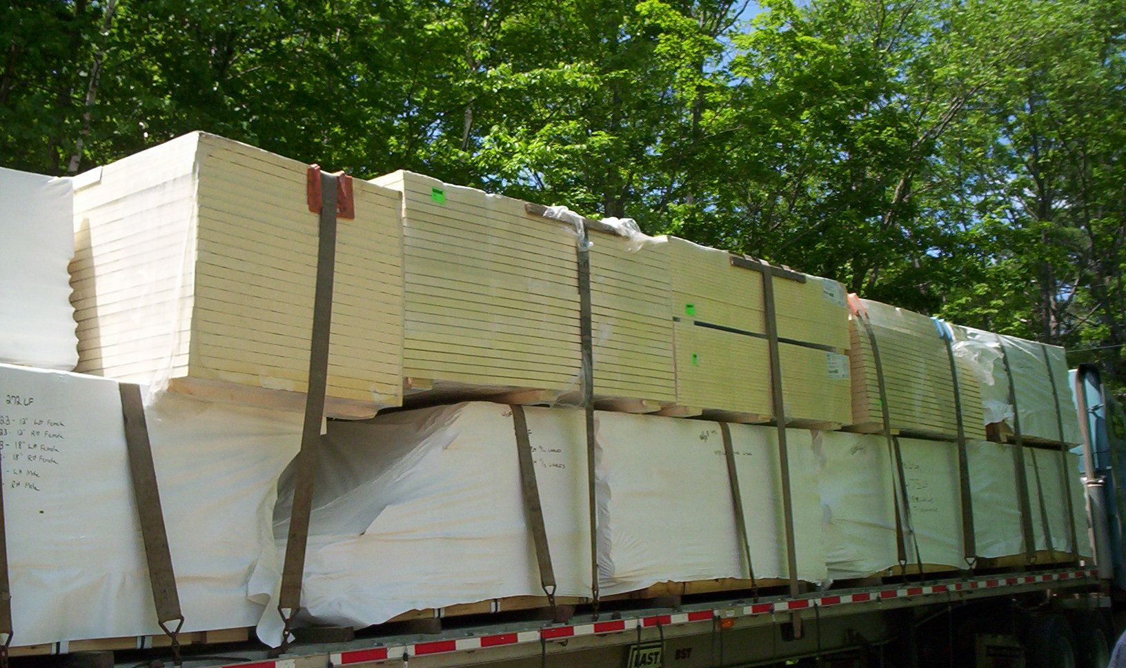 Loaded flatbed truck stacked with white and beige wrapped cargo outdoors under trees