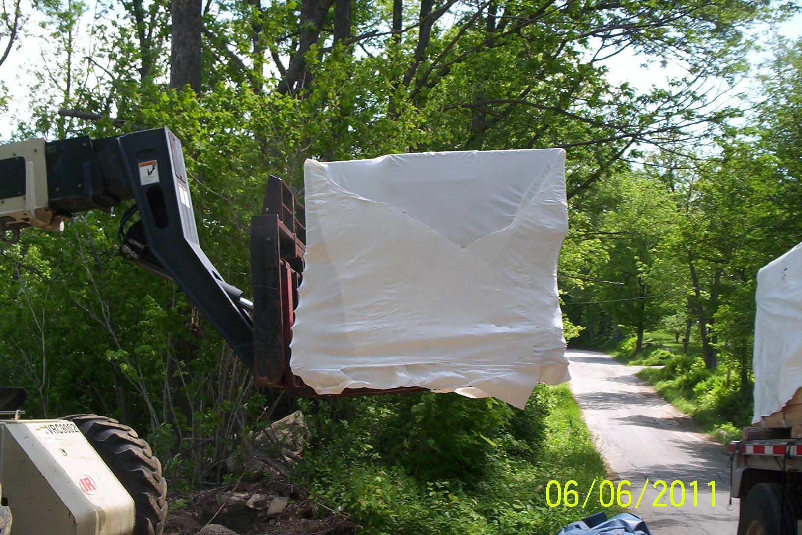 Excavator lifting a large white wrapped bundle beside a wooded dirt road.