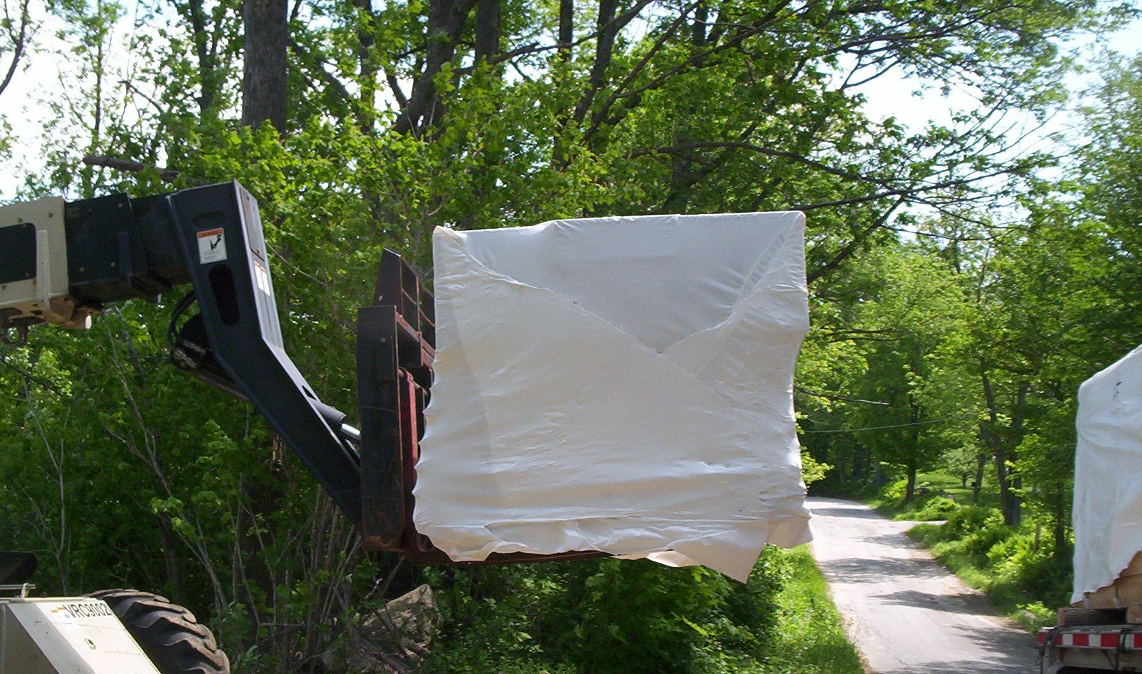 Forklift carrying a large white-wrapped pallet outdoors beside a tree-lined path