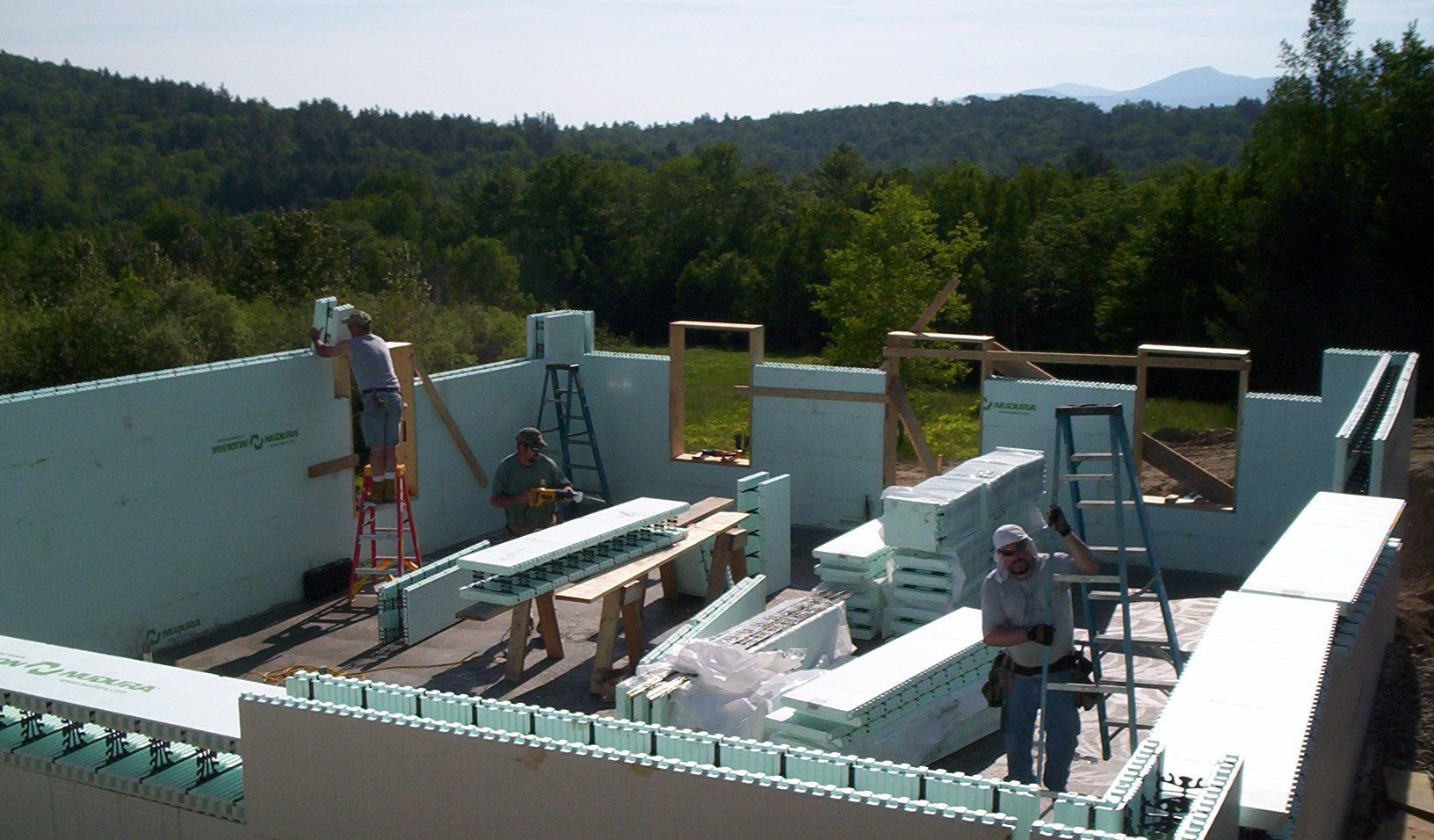 Workers installing white walls on a rooftop under construction, with forested hills in the background