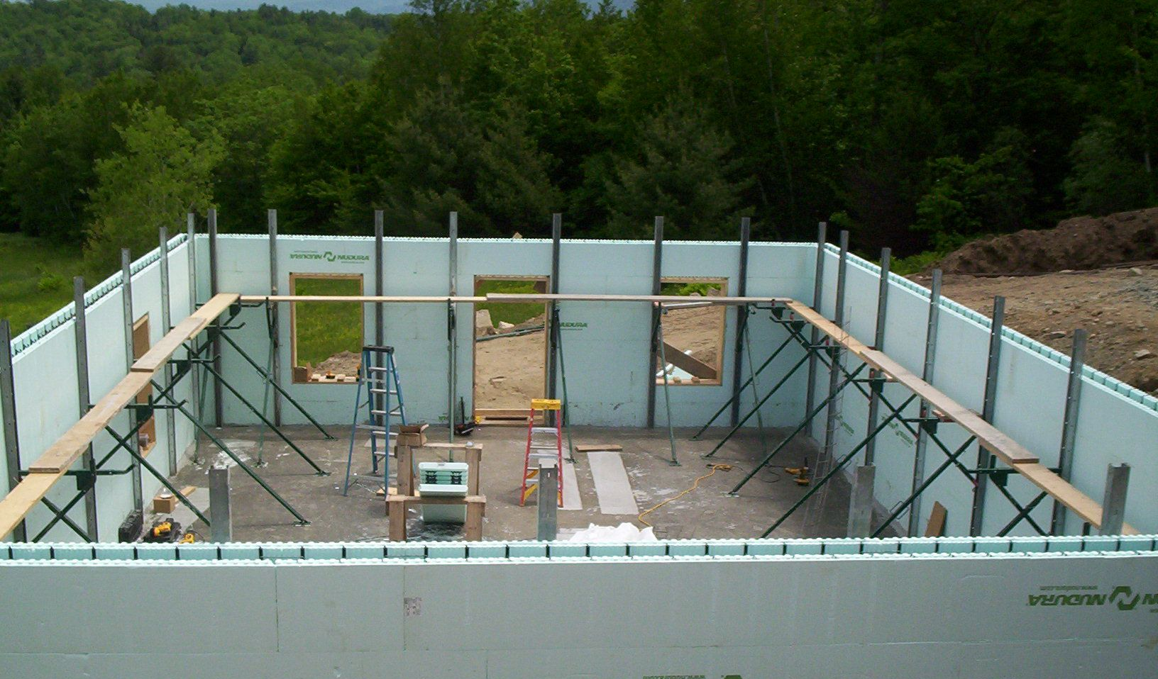 Partially built foundation with white insulated walls, scaffolding, and a wooded background