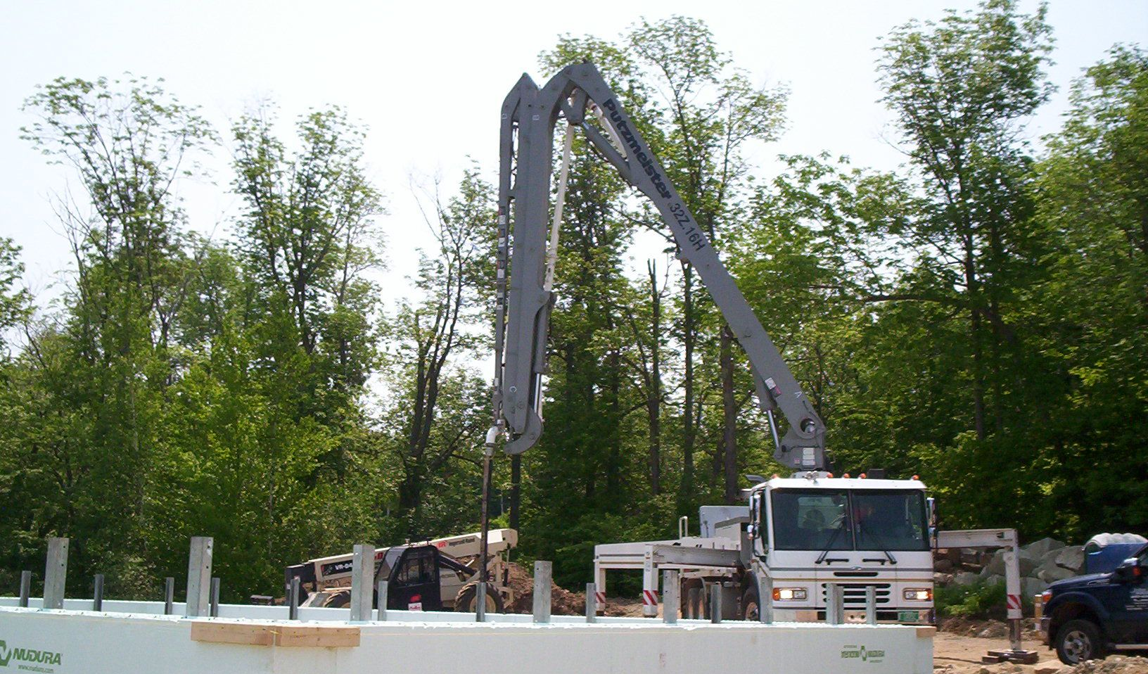 Construction crane lifting materials near a white truck at a roadside work site with trees in the background