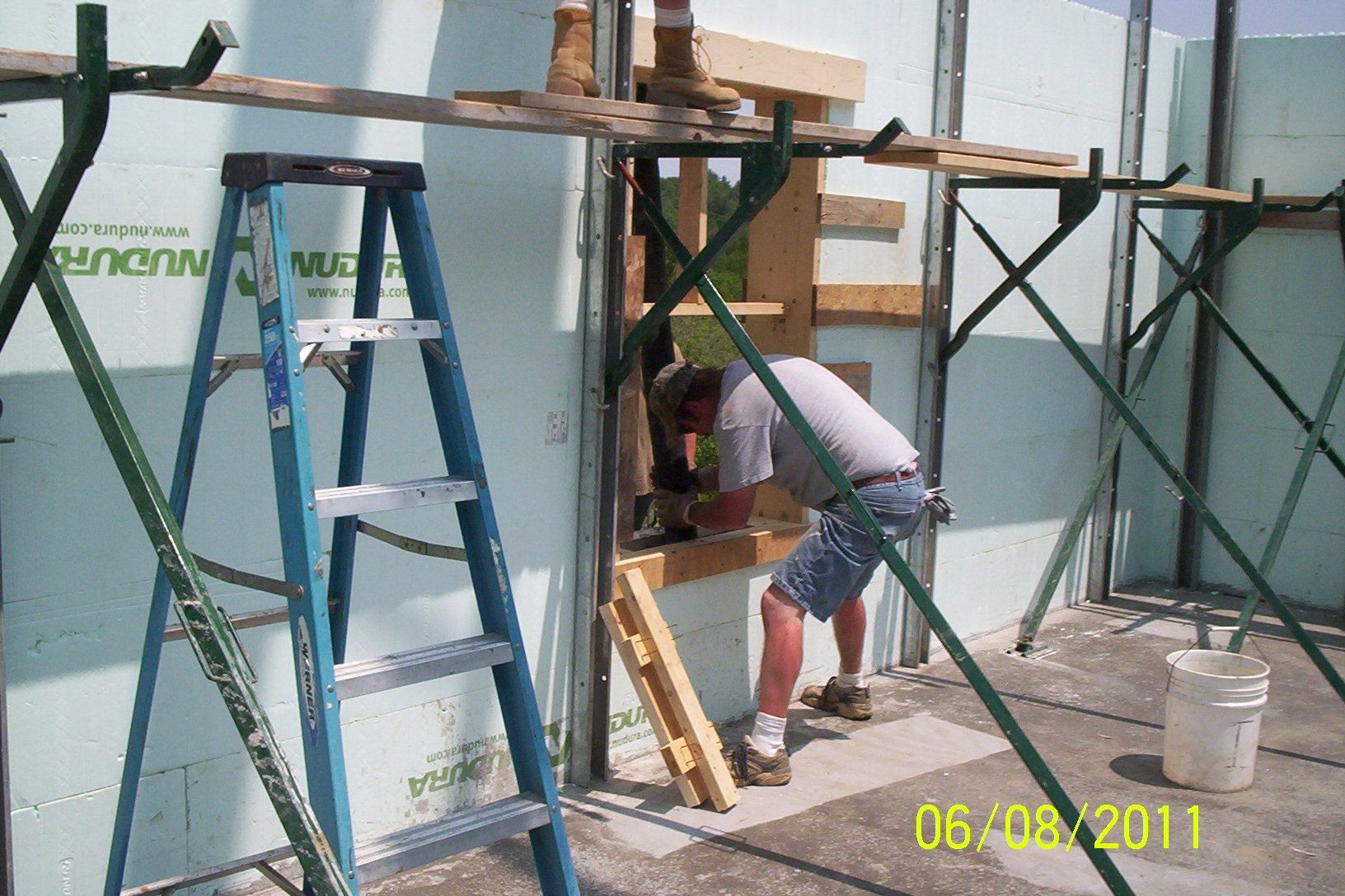 Worker bending into a wall opening on a construction site, with ladders and scaffolding nearby.