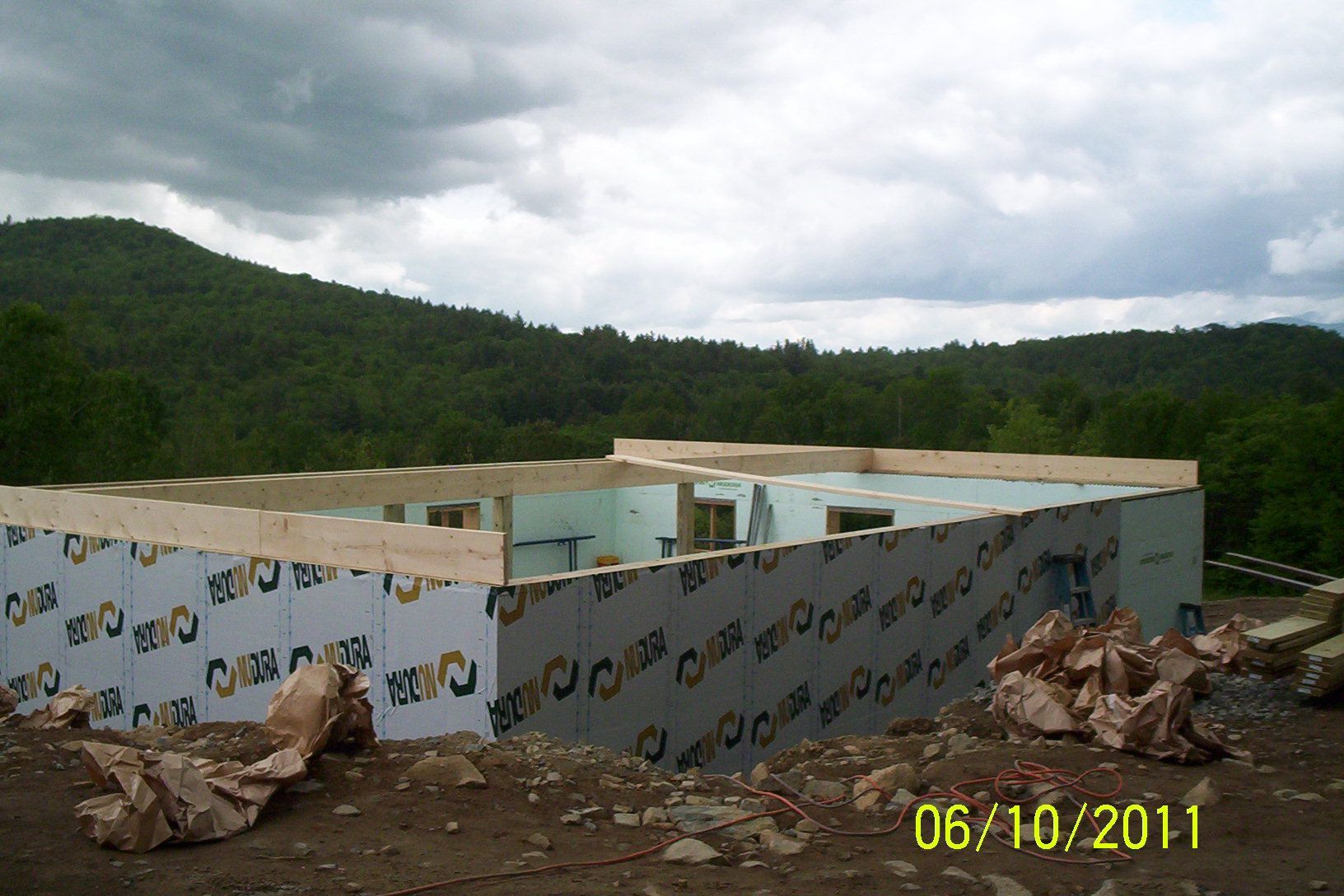 Partially built building with blue insulation panels in a forested mountain area under cloudy skies