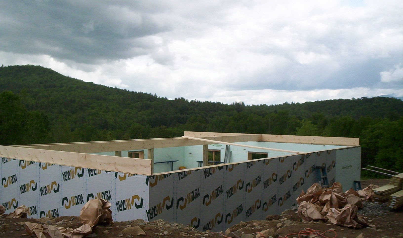 A low industrial building under construction amid debris, with forested hills and cloudy sky in the background