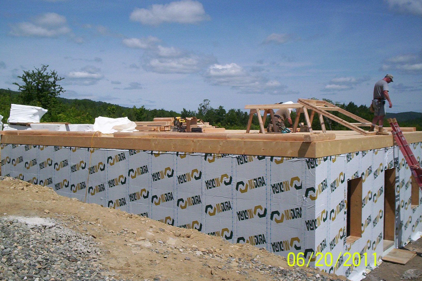 Partially built house foundation wrapped in white insulation, with wooden framing and a worker on the roof.