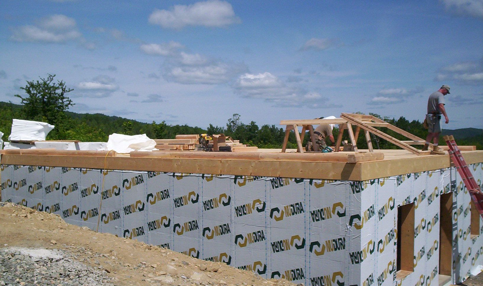 Construction site with unfinished wood roof framing atop an insulated foundation under a blue sky
