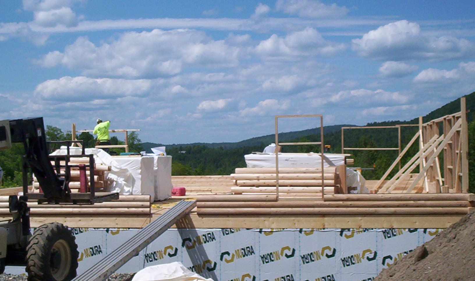 Construction materials on a flatbed truck at a site under a cloudy sky, with stacked lumber and white bundles.