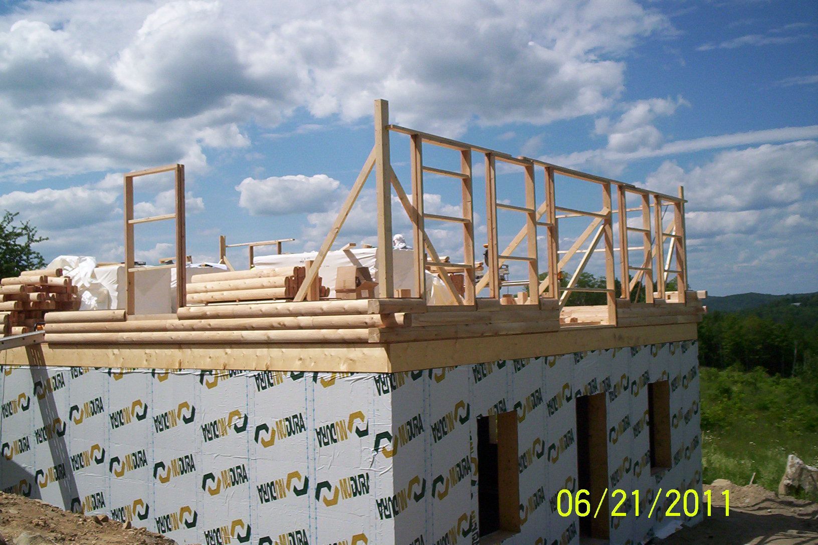 Wood-frame house under construction with a wooden roof frame atop wrapped exterior walls under a partly cloudy sky