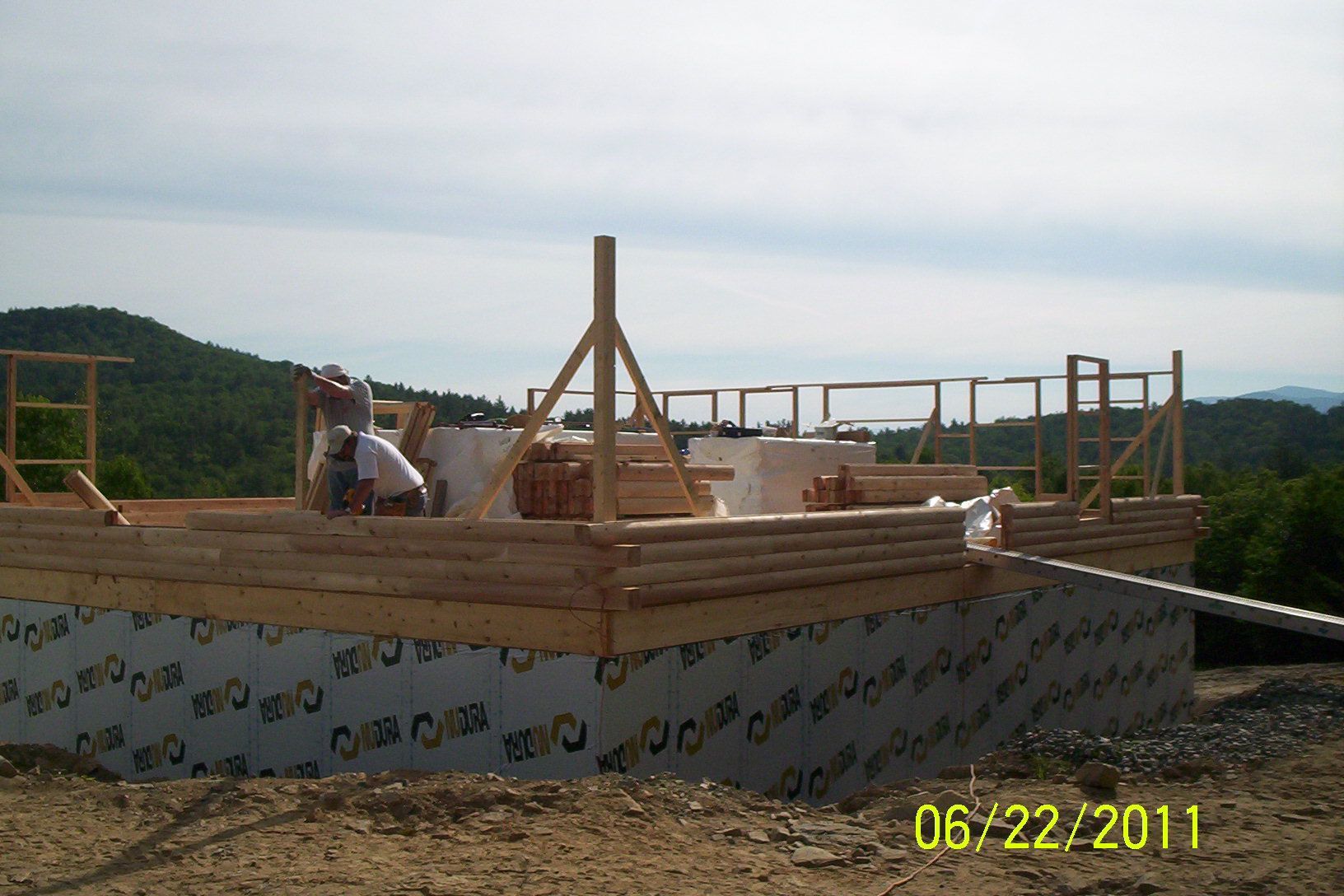 Wooden house frame under construction on a foundation, with distant hills and a date stamp.