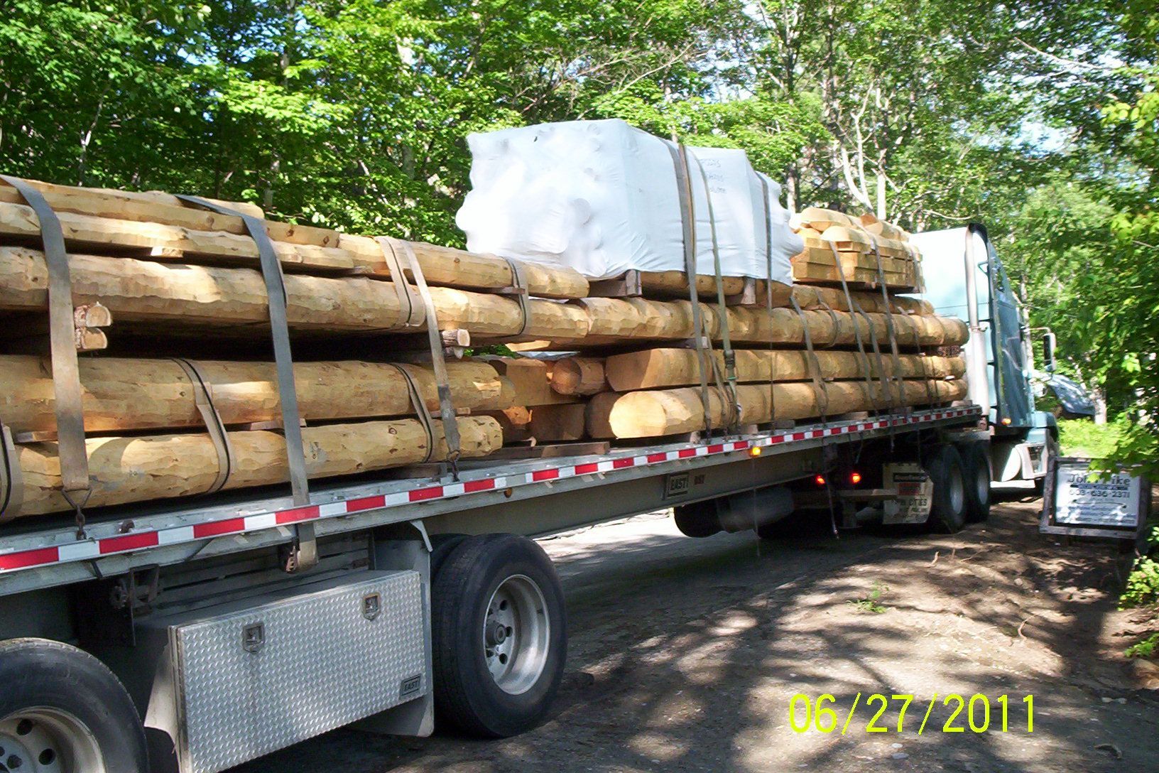 Flatbed truck loaded with stacked logs and a wrapped white bundle on a tree-lined road