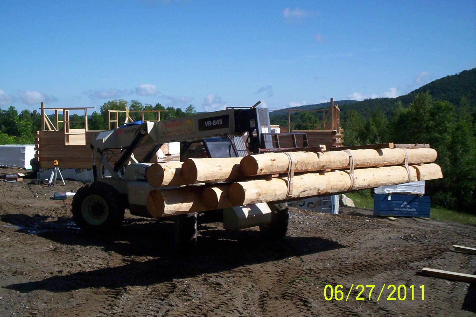 Logging truck loaded with cut timber on a dirt lot, with mountains in the background.